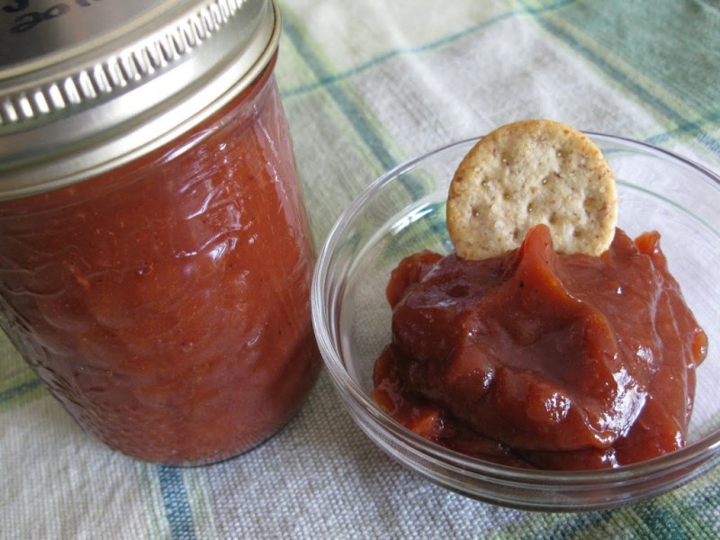 A jar of apple butter beside a small bowl with a cracker dipping into apple butter.