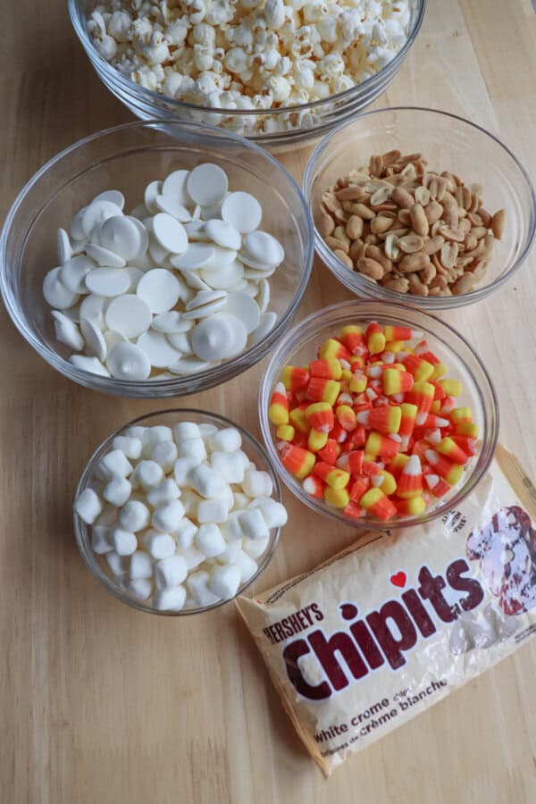 Ingredients for candy corn popcorn balls in clear glass bowls on a wooden table.