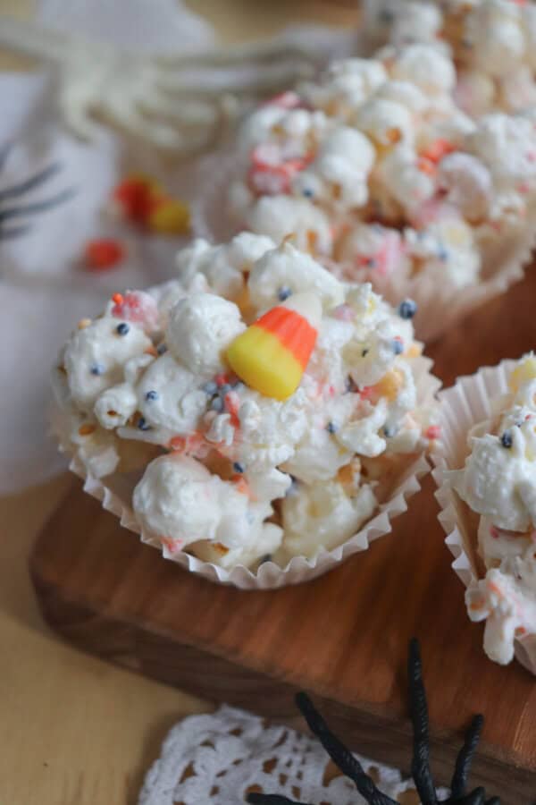 Popcorn balls with Halloween sprinkles and candy corn in cupcake liners on a wooden cutting board.