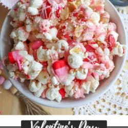 A bowl of Valentines Day themed popcorn snack mix, featuring popcorn drizzled with pink and red icing, heart-shaped candies, and sprinkles. A lacy napkin and pink cupcake liners sit nearby, adding charm to the scene. Text reads: Valentines Day Popcorn Snack Mix with chocolate coconut date bars.”.