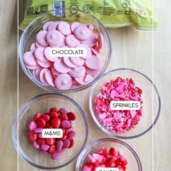 Ingredients for a treat mix on a table: a bag of sea salt popcorn, a bowl of pink chocolate wafers, chocolate coconut date bars, colorful sprinkles, red and pink M&Ms, and heart-shaped candies.