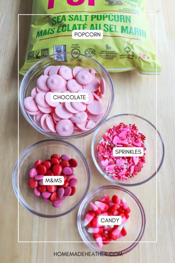 Ingredients for a treat mix on a table: a bag of sea salt popcorn, a bowl of pink chocolate wafers, chocolate coconut date bars, colorful sprinkles, red and pink M&Ms, and heart-shaped candies.