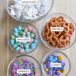 Top view of a table with five bowls containing different snacks: white chocolate wafers, pretzels, candy-coated mini eggs, colorful sprinkles, and round purple and yellow candies. Nearby sits a plate of inviting chocolate coconut date bars. Each bowl is labeled accordingly.
