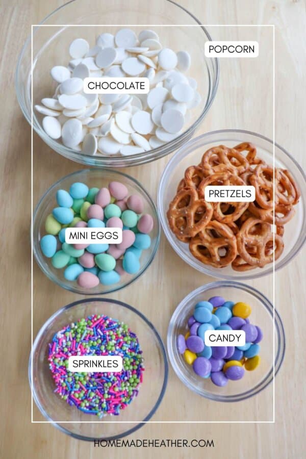 Top view of a table with five bowls containing different snacks: white chocolate wafers, pretzels, candy-coated mini eggs, colorful sprinkles, and round purple and yellow candies. Nearby sits a plate of inviting chocolate coconut date bars. Each bowl is labeled accordingly.