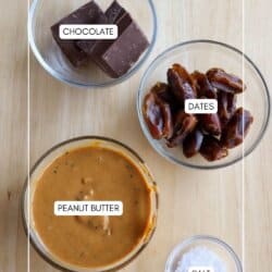 Top view of four glass bowls on a wooden surface containing ingredients: chocolate, dates, peanut butter mixed with chia seeds, and salt, perfect for creating delicious date bites.