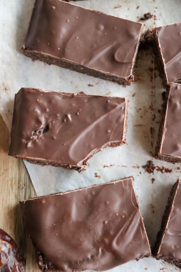 Close-up of chocolate coconut date bars on parchment paper, with one bar partially eaten. The bars have a smooth, glossy surface and are arranged in rows, showcasing their rich, dark color.