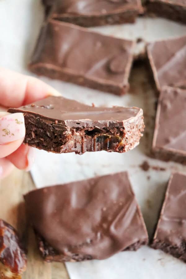 A hand holding a square piece of chocolate coconut fudge with a bite taken out of it. The background shows more pieces on parchment paper, some overlapping. The fudge has a smooth, glossy surface reminiscent of decadent date bars.