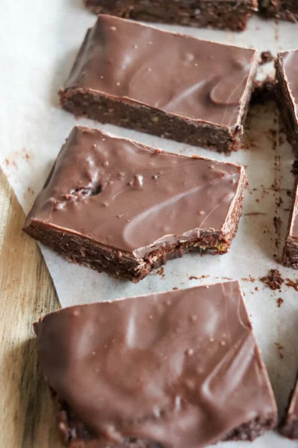 Three rectangular chocolate brownies rest on parchment paper, topped with a smooth, glossy chocolate finish. One brownie has a bite taken out of it, revealing the dense, moist texture reminiscent of rich chocolate coconut date bars. They are placed on a wooden surface.
