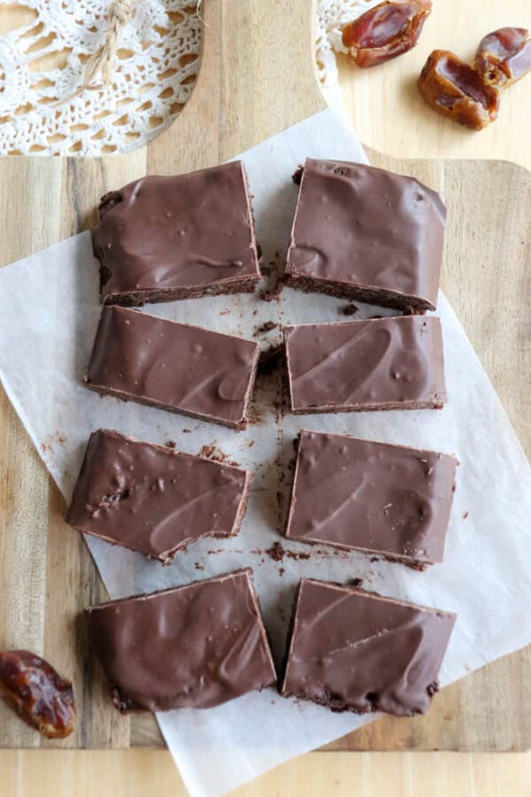 Eight rectangular chocolate coconut date bars are arranged on parchment paper on a wooden cutting board. Some whole dates are scattered around, and a doily peeks into the top corner of the image.