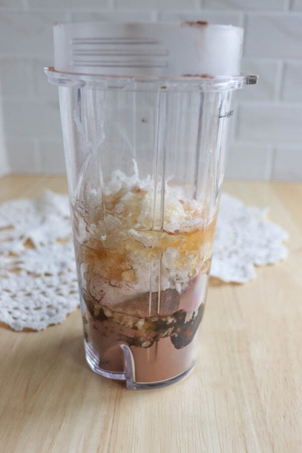 A clear blender cup filled with various ingredients, including ice, banana pieces, and a brown powder—possibly cocoa or coffee—awaits transformation into a delicious treat. Nearby, chocolate coconut date bars rest on the wooden table adorned with a white lace doily in the background.