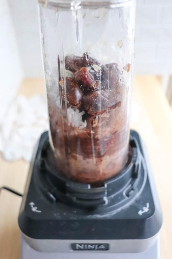 Close-up of a blender containing ice cubes, cherries, and other ingredients ready to turn into a delicious mix. A hint of chocolate coconut date bars completes the scene. The transparent container sits on a black base, with a softly blurred background bathed in natural light.