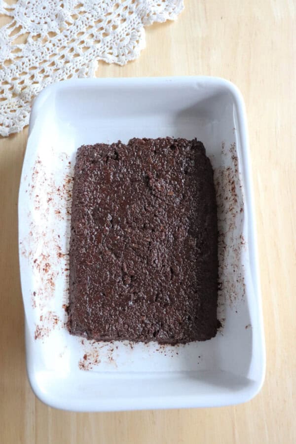 A rectangular white baking dish holds a dense chocolate cake adorned with hints of coconut, resting on a wooden surface. A crocheted lace piece peeks from the corner, adding a touch of charm.