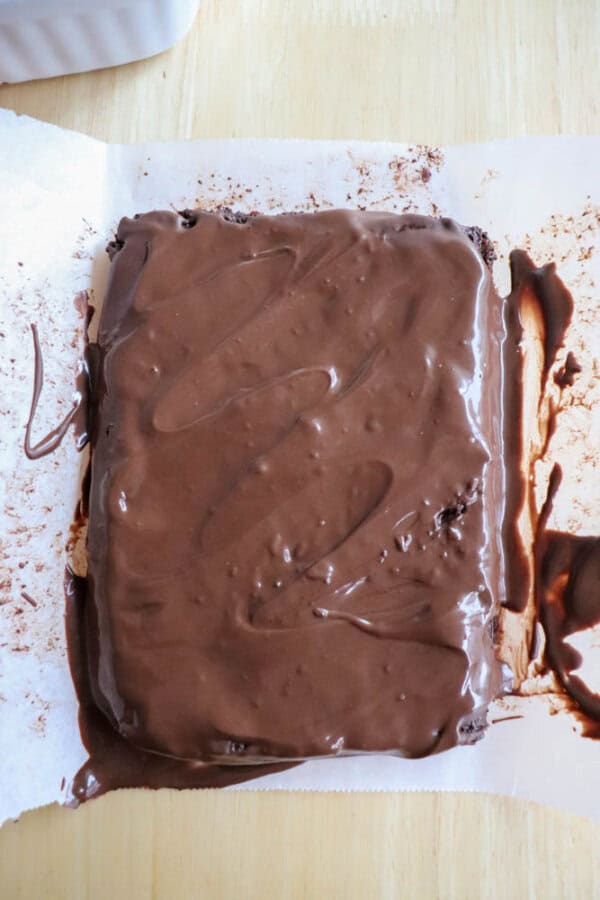 A thick slab of chocolate fudge lies on parchment paper, partially coated with a layer of melted chocolate. Some has spilled over the edges, creating a messy, appetizing appearance reminiscent of delightful chocolate coconut date bars on a light wooden surface.