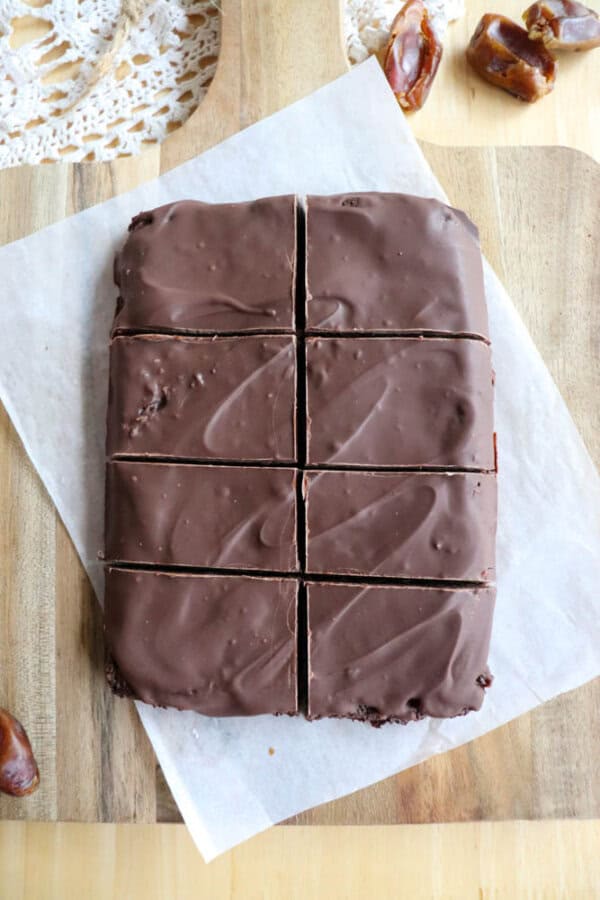 Eight square chocolate coconut date bars are arranged on a piece of parchment paper, placed on a wooden cutting board. Whole dates are scattered around, hinting they might be an ingredient. A lace doily is partially visible in the background.
