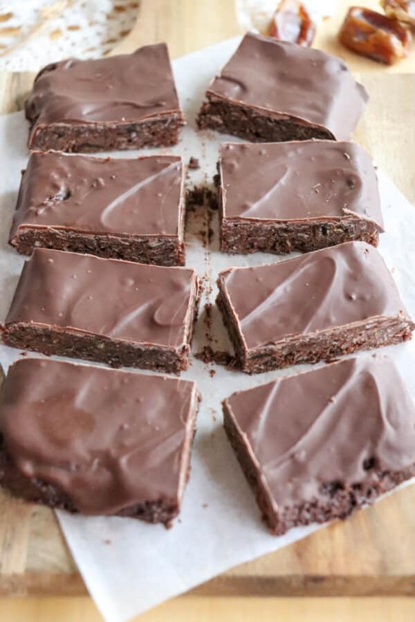 Eight rectangular chocolate-covered brownies are arranged on a piece of parchment paper on a wooden surface. The smooth, glossy coating is evenly spread atop each chocolate coconut date bar, creating a tempting and delicious appearance.