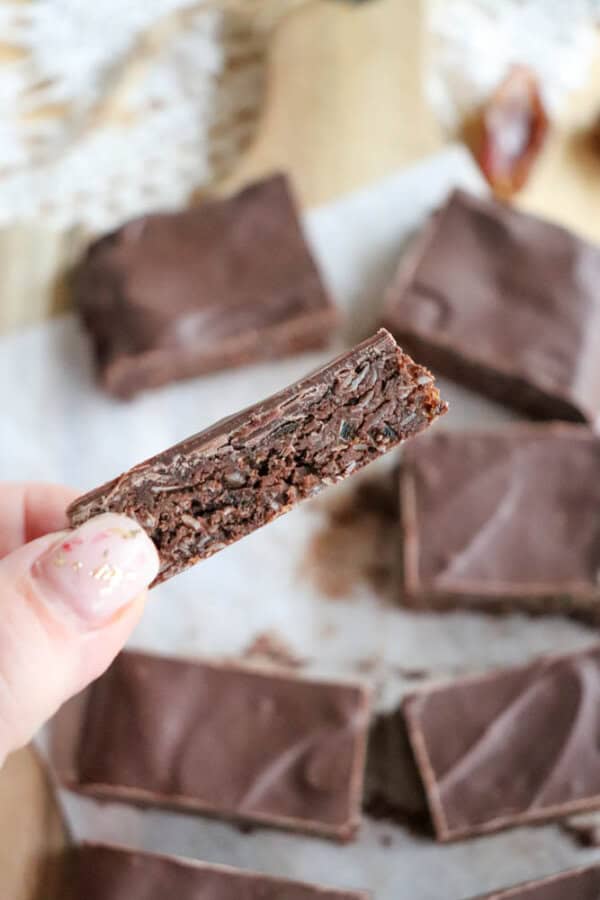A hand holds a chocolate brownie with a bite taken out of it, reminiscent of the indulgent texture found in chocolate coconut date bars. Several more brownies are elegantly arranged on parchment paper, showcasing their rich, dark appearance with a glossy chocolate top layer.