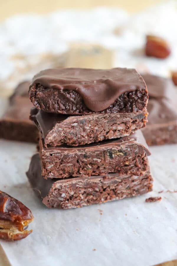 A stack of chocolate coconut date bars sits on parchment paper. The bars have a rich, textured appearance, topped with a smooth layer of chocolate. Dates are scattered around as decoration.