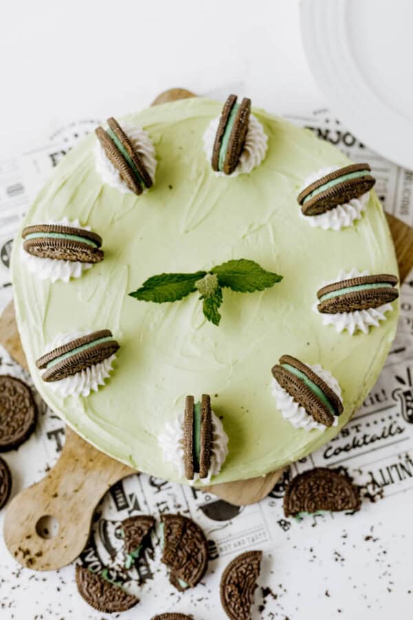 A round Mint Oreo No Bake Cheesecake with a mint green hue, topped with six whipped cream dollops, each crowned by a mint chocolate sandwich cookie and fresh mint. Cookie crumbs are scattered around the cake.