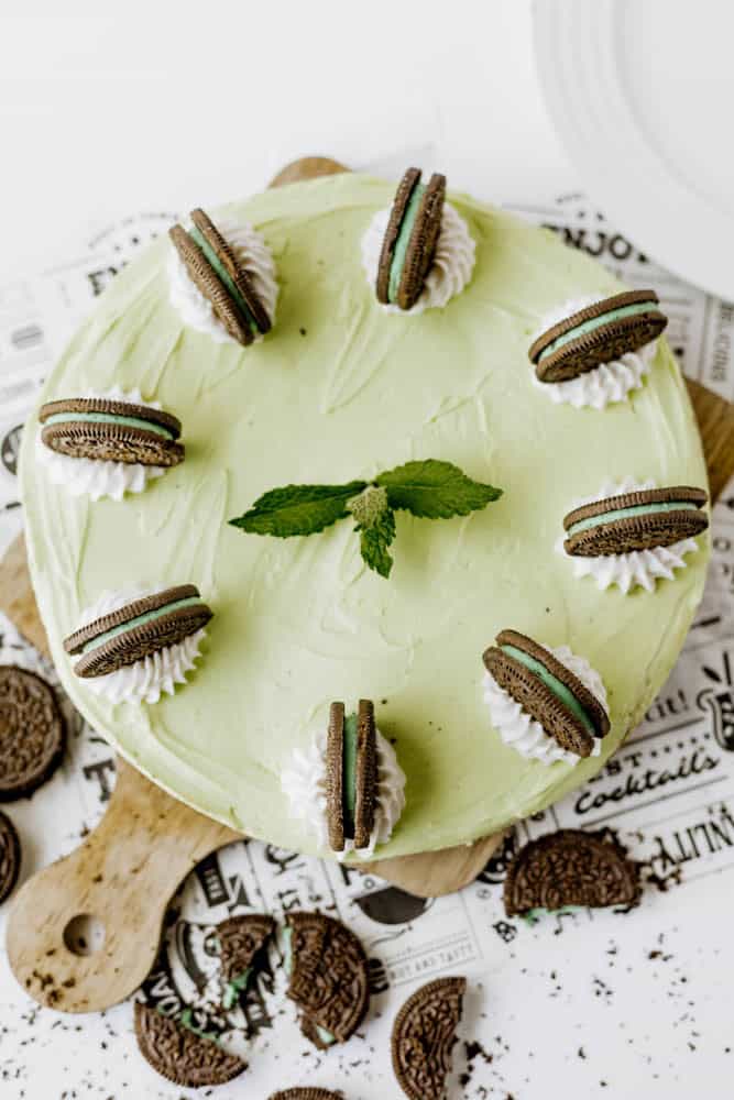 A round Mint Oreo No Bake Cheesecake with a mint green hue, topped with six whipped cream dollops, each crowned by a mint chocolate sandwich cookie and fresh mint. Cookie crumbs are scattered around the cake.