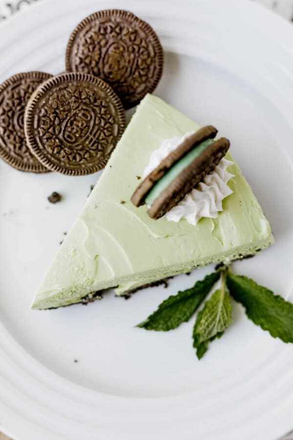 A slice of light green Mint Oreo No Bake Cheesecake on a white plate, topped with whipped cream and a chocolate sandwich cookie, garnished with mint leaves and more cookies on the side.