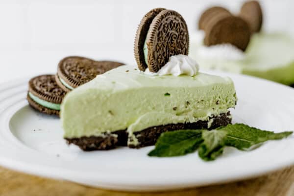A slice of Mint Oreo No Bake Cheesecake sits on a white plate, topped with whipped cream and an Oreo cookie, with extra cookies and fresh mint leaves as garnish.