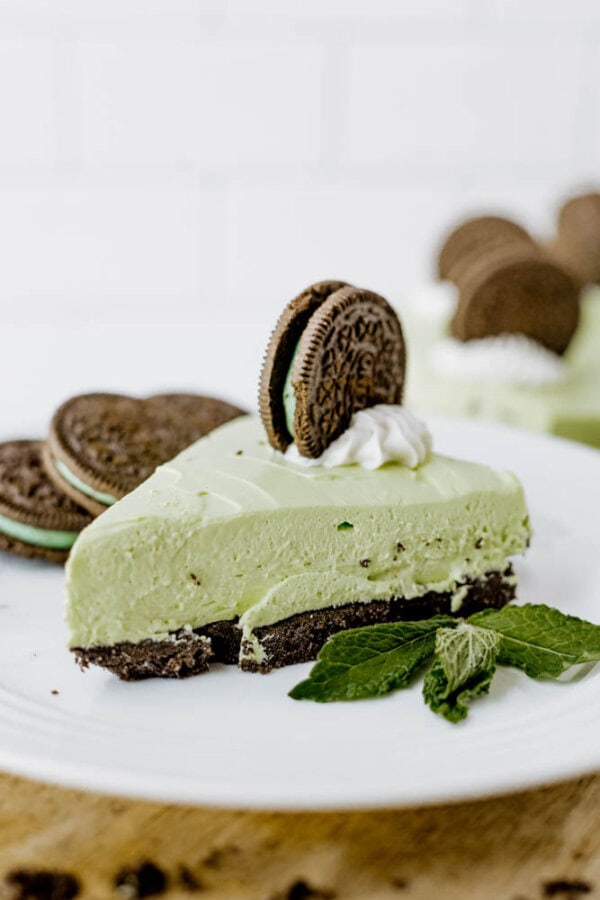 A slice of Mint Oreo No Bake Cheesecake with an Oreo cookie crust, topped with an Oreo cookie and a dollop of whipped cream, garnished with fresh mint leaves on a white plate.
