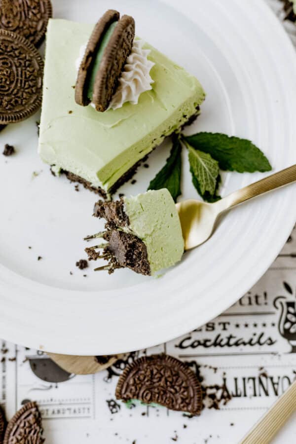 A slice of Mint Oreo No Bake Cheesecake is topped with a chocolate sandwich cookie and whipped cream on a white plate, with a gold fork holding a bite. Crumbled cookies and mint leaves complete the tempting presentation.