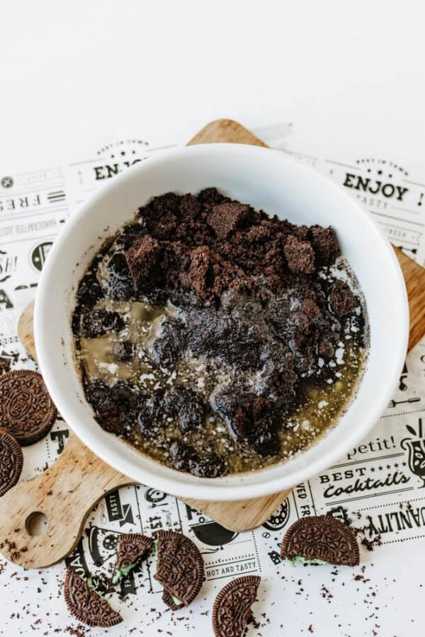 A white bowl filled with crumbled Mint Oreo No Bake Cheesecake crust—crushed cookies blended with melted butter—sits on a decorative paper atop a wooden board, with more cookie pieces scattered around.