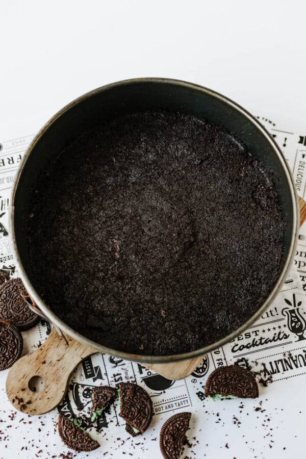 A round baking pan filled with a dark, crumbly chocolate cookie crust sits on a wooden board, surrounded by broken cookies and crumbs—perfect as the base for a Mint Oreo No Bake Cheesecake.