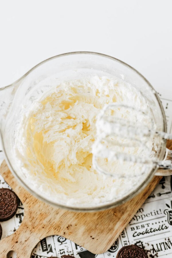 A glass bowl filled with fluffy whipped cream sits on a wooden board, ready for a Mint Oreo No Bake Cheesecake. An electric mixer coated in cream rests nearby, with chocolate sandwich cookies partially visible at the edge of the image.