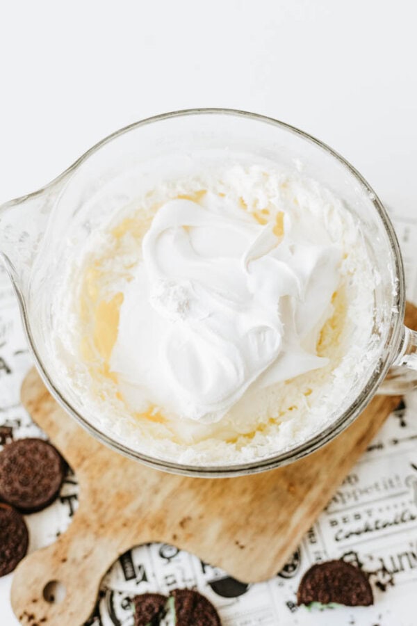A glass mixing bowl filled with a creamy Mint Oreo No Bake Cheesecake mixture sits on a wooden board, with chocolate sandwich cookies nearby on a printed cloth.