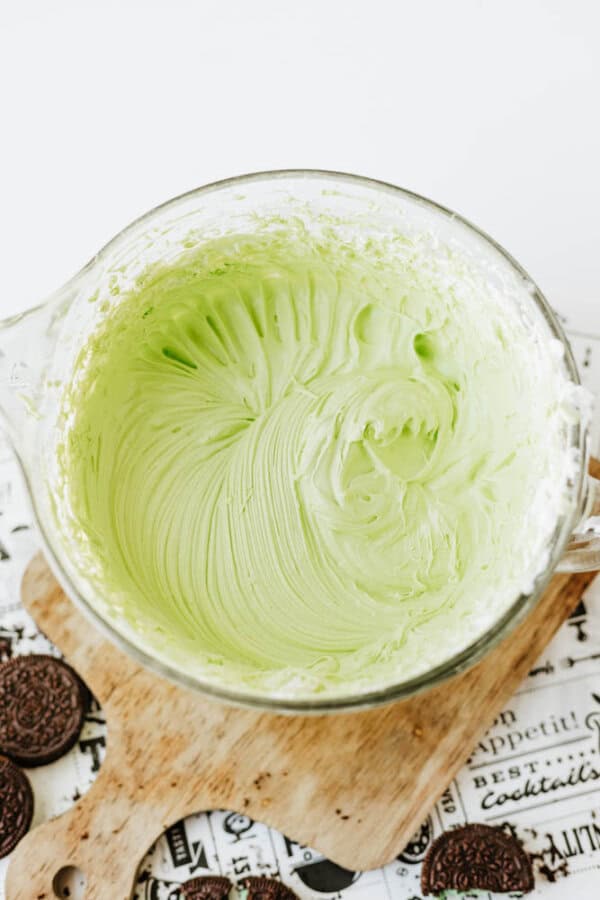 A glass bowl filled with creamy, light green frosting for Mint Oreo No Bake Cheesecake sits on a wooden cutting board, with chocolate sandwich cookies partially visible nearby.