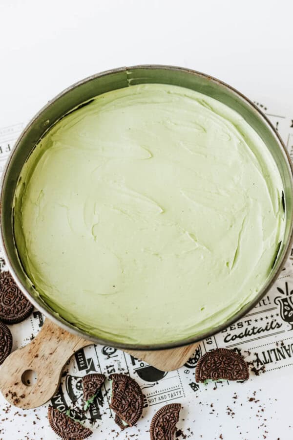 A round pan filled with smooth, light green Mint Oreo No Bake Cheesecake rests on a wooden board, surrounded by crumbled chocolate sandwich cookies on a decorative paper.