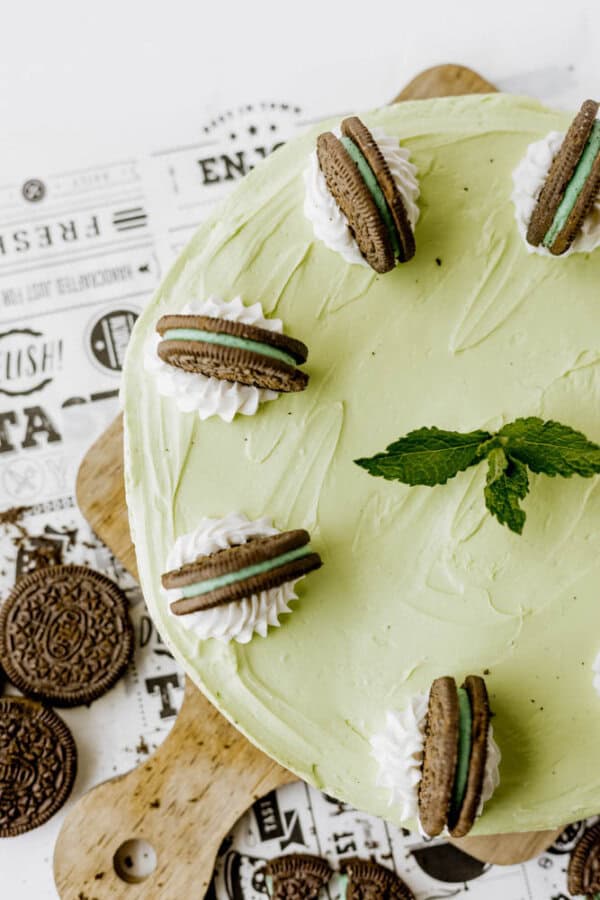A round Mint Oreo No Bake Cheesecake in mint green, topped with whipped cream, chocolate sandwich cookies, and a sprig of fresh mint, displayed on a wooden board with extra cookies on the side.