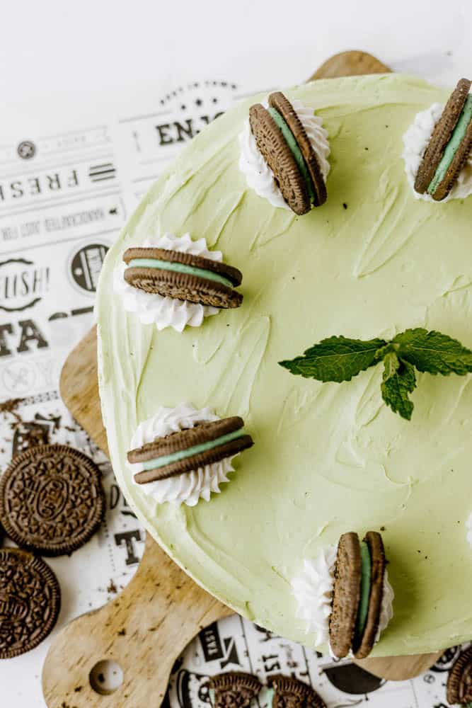 A round Mint Oreo No Bake Cheesecake in mint green, topped with whipped cream, chocolate sandwich cookies, and a sprig of fresh mint, displayed on a wooden board with extra cookies on the side.