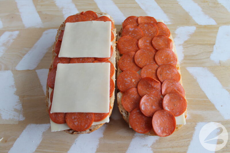 French loaf of bread cut in half and laid open with pizza sauce, pepperoni slices and mozzarella cheese slices covering it.
