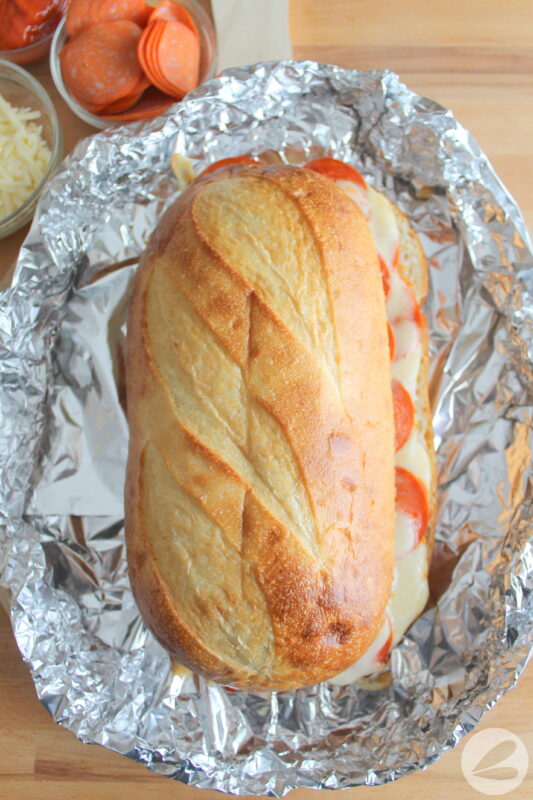 Large loaf of bread with pepperoni and melted cheese oozing out the sides on a bed of foil.