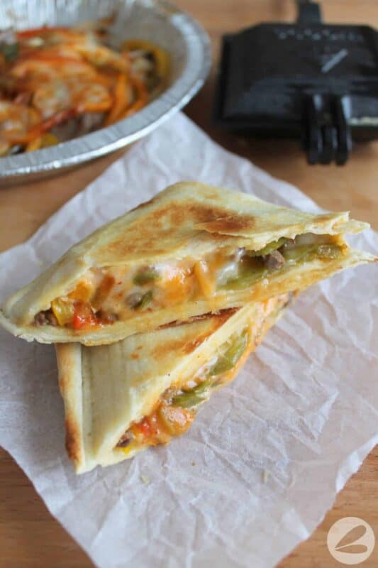 Camp cooker fajita cut in half diagonally to show vegetables, steak and melty cheese inside, on a piece of parchment paper with a pie iron in the background.
