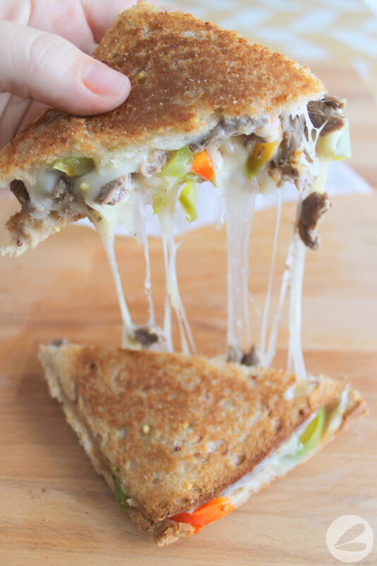 Pie Iron Philly Cheesesteak Sandwich cut in half diagonally to show filling, with one half held in hand and pulled with cheese pulling from both halves.