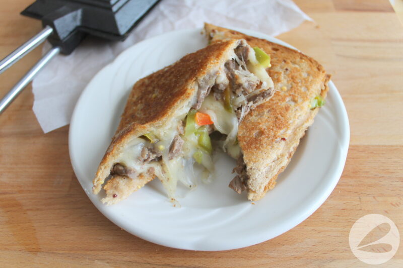 Pie Iron Philly Cheesesteak Sandwich cut in half diagonally to show filling.  Cooked steak and bell peppers ooze out with white melty cheese.