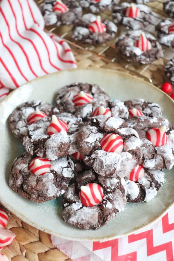 A plate of chocolate cookies dusted with powdered sugar and adorned with red and white striped Hershey's Kisses evokes the charm of candy cane brownie blossoms. More cookies cool on a wire rack in the background, set on a woven surface with a red and white striped cloth nearby.