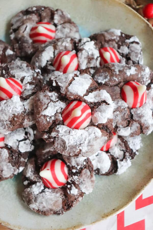 A plate of chocolate crinkle cookies topped with red and white striped Hershey's Kisses, resembling candy cane brownie blossoms. The cookies are dusted with powdered sugar, creating a festive and inviting appearance.
