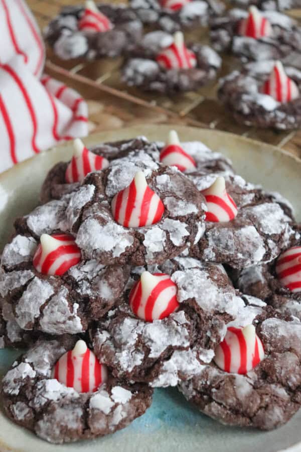 A plate of crinkle chocolate cookies, resembling candy cane brownie blossoms, topped with red and white striped candy kisses, dusted with powdered sugar. More cookies are visible on a cooling rack in the background, alongside a red and white striped cloth.