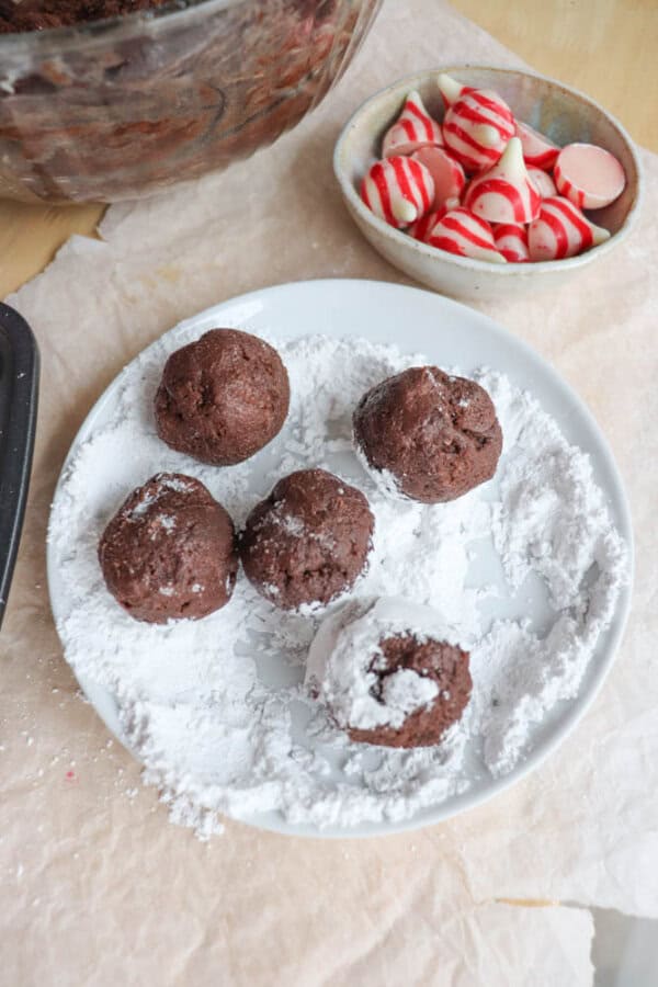 Chocolate cookie dough balls are being rolled in powdered sugar on a white plate, resembling candy cane brownie blossoms. Nearby, there is a bowl with red and white striped chocolate candies. A mixing bowl with additional dough is partially visible.