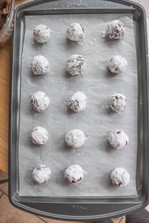 A baking tray lined with parchment paper holds fifteen round chocolate dough balls coated in powdered sugar, like little candy cane delights, evenly spaced in three columns of five.