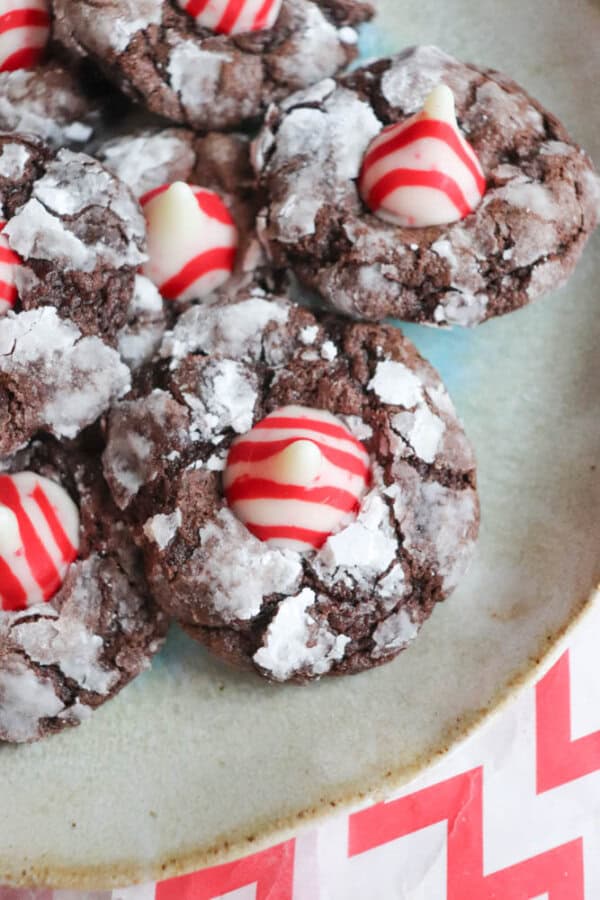 A plate of chocolate crinkle cookies topped with red and white striped Hershey's Kisses resembles candy cane brownie blossoms. The cookies, dusted with powdered sugar, are arranged in a pile on a table adorned with a red and white chevron-patterned cloth.