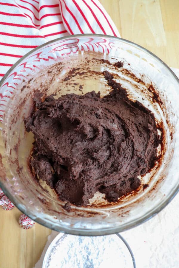 A glass bowl filled with thick, dark chocolate cookie dough for candy cane brownie blossoms sits on a wooden countertop. A red and white striped cloth is partially visible in the background. A small sifter with powdered sugar is nearby.