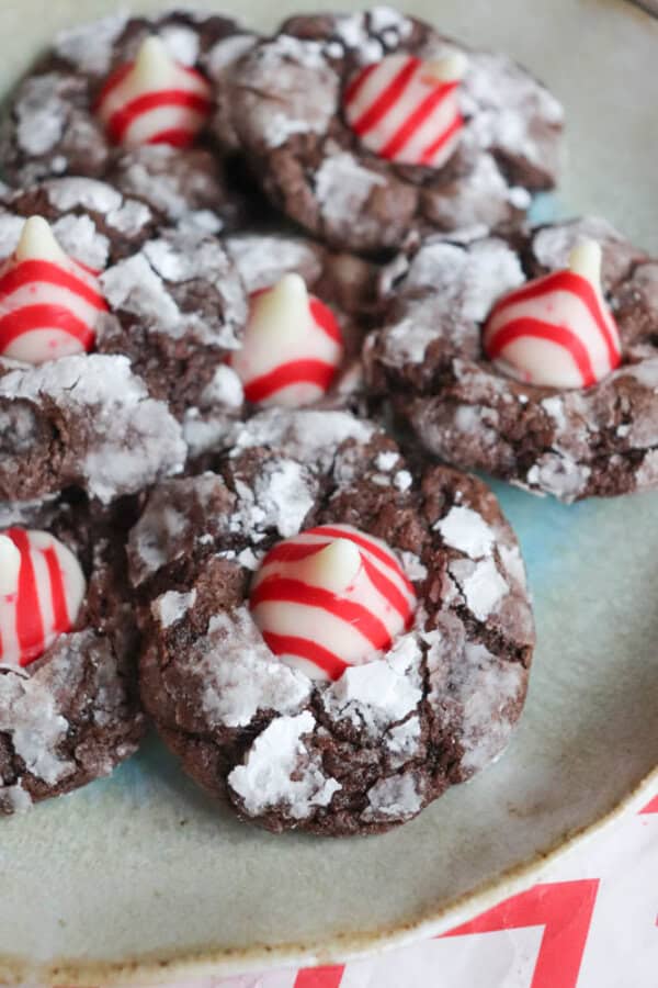 A plate of candy cane brownie blossom cookies topped with red and white striped Hershey's Kisses, dusted with powdered sugar. The cookies are arranged closely together on a round plate.