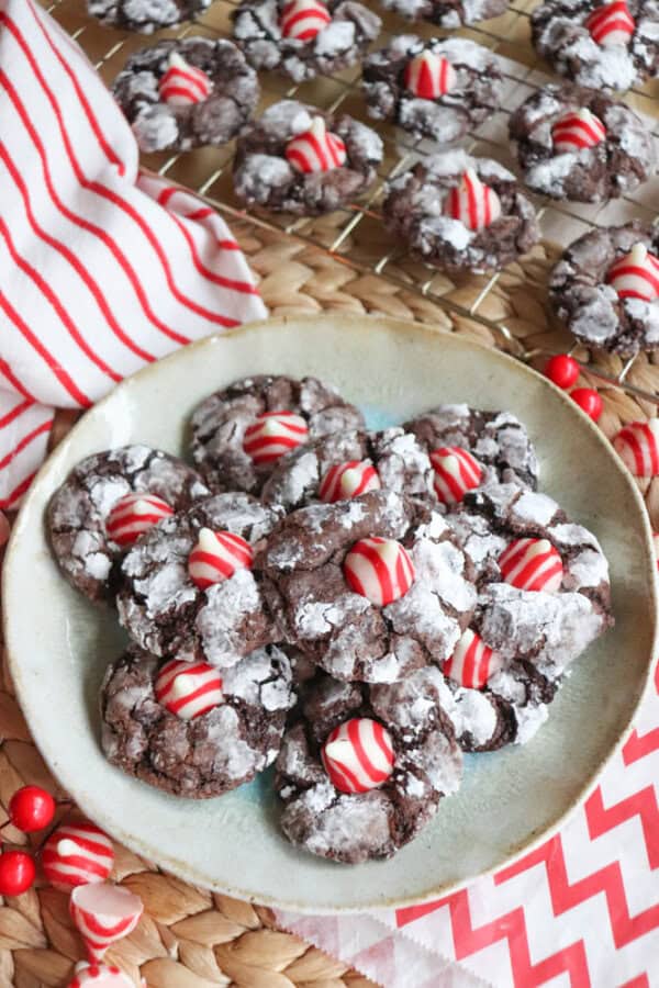 A plate of chocolate crinkle cookies topped with red and white striped candies reminiscent of candy cane brownie blossoms. More cookies cool on a wire rack behind, surrounded by festive striped napkins and red candies, creating a delightful holiday scene.