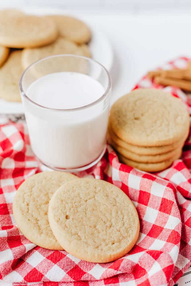 A glass of milk and several sugar cookies rest on a red and white checkered cloth, evoking the coziness of a homemade Snickerdoodle Cookie recipe, with more cookies on a plate in the background.